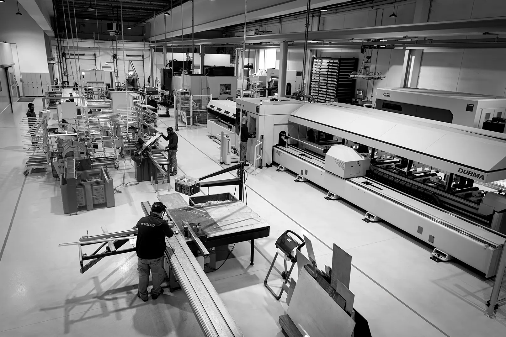 Homem operando máquina industrial em fábrica de metal com várias máquinas e trabalhadores.