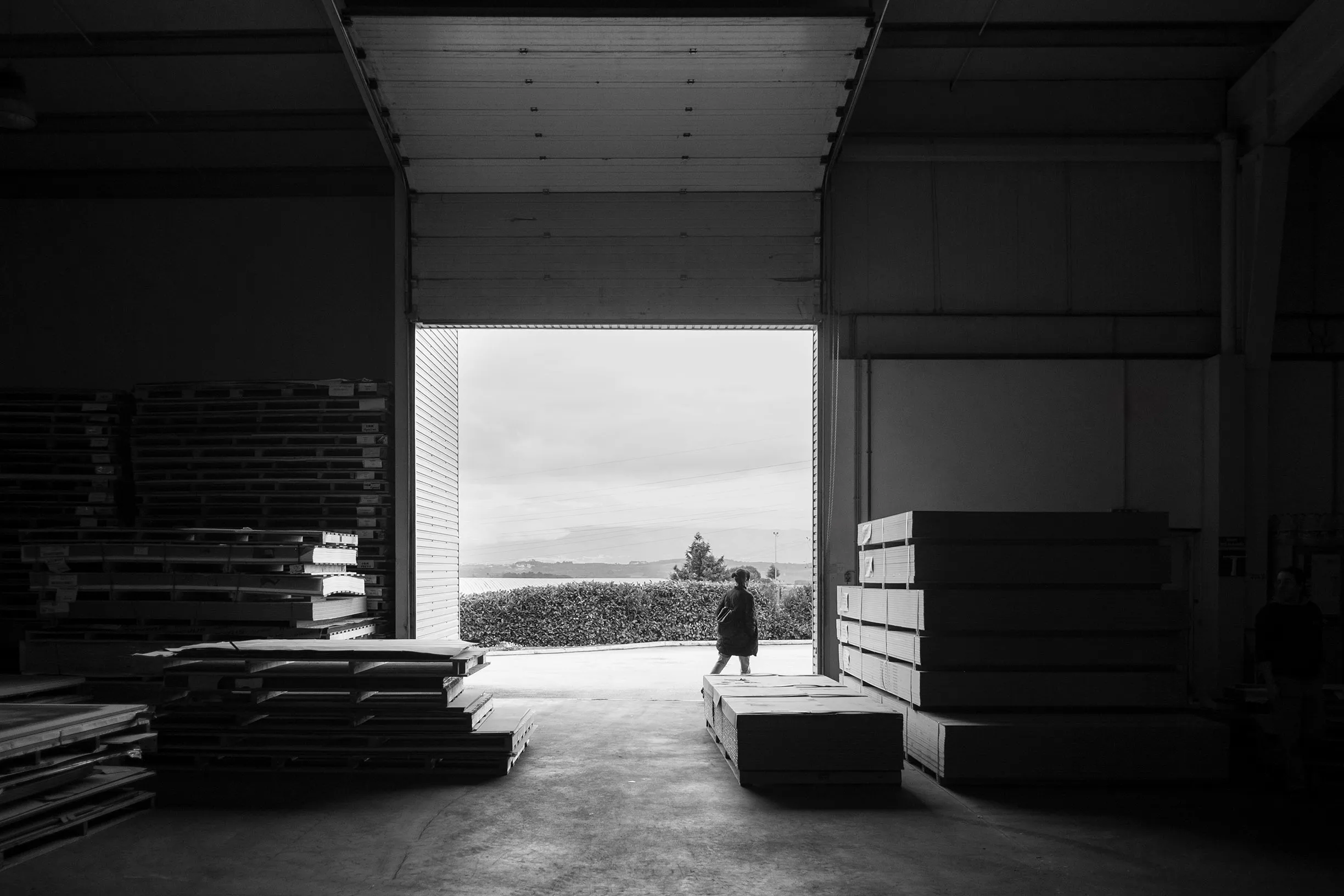 Interior de armazém escuro com pilhas de madeira e pessoa caminhando na porta aberta.