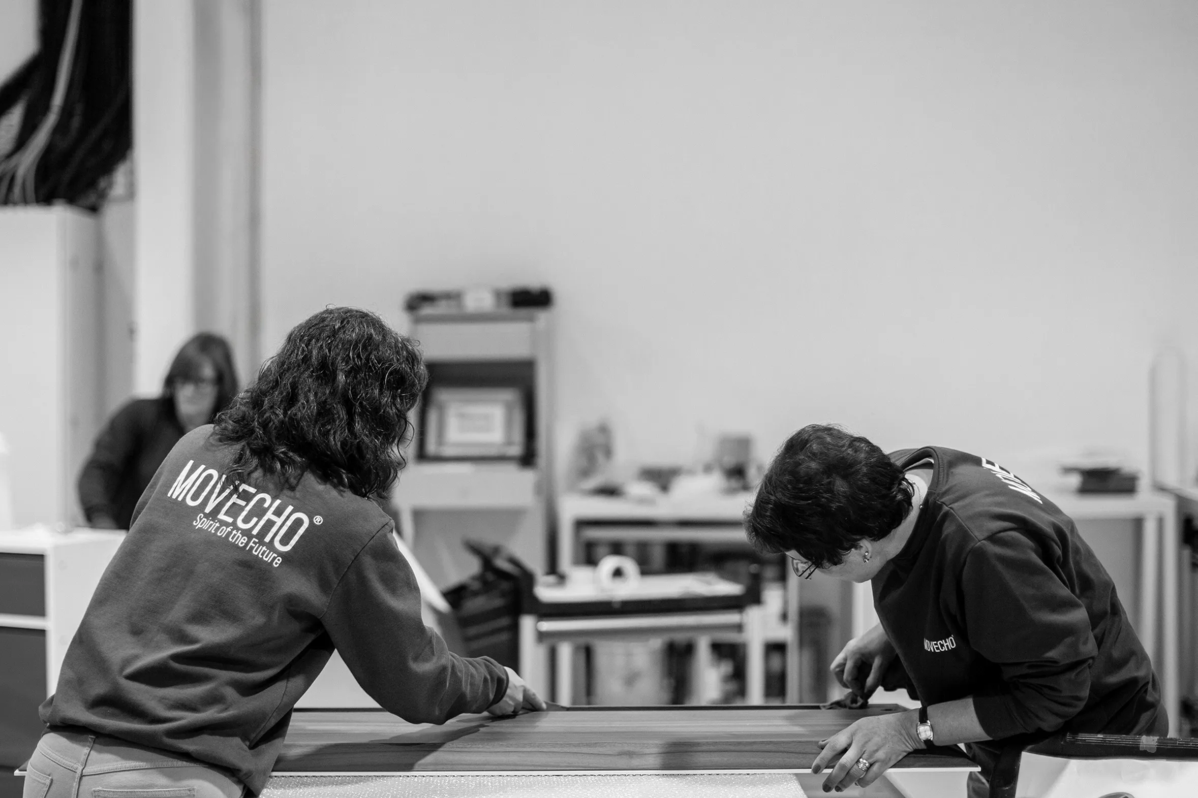 Duas mulheres medem cuidadosamente uma peça de madeira dentro de uma oficina.