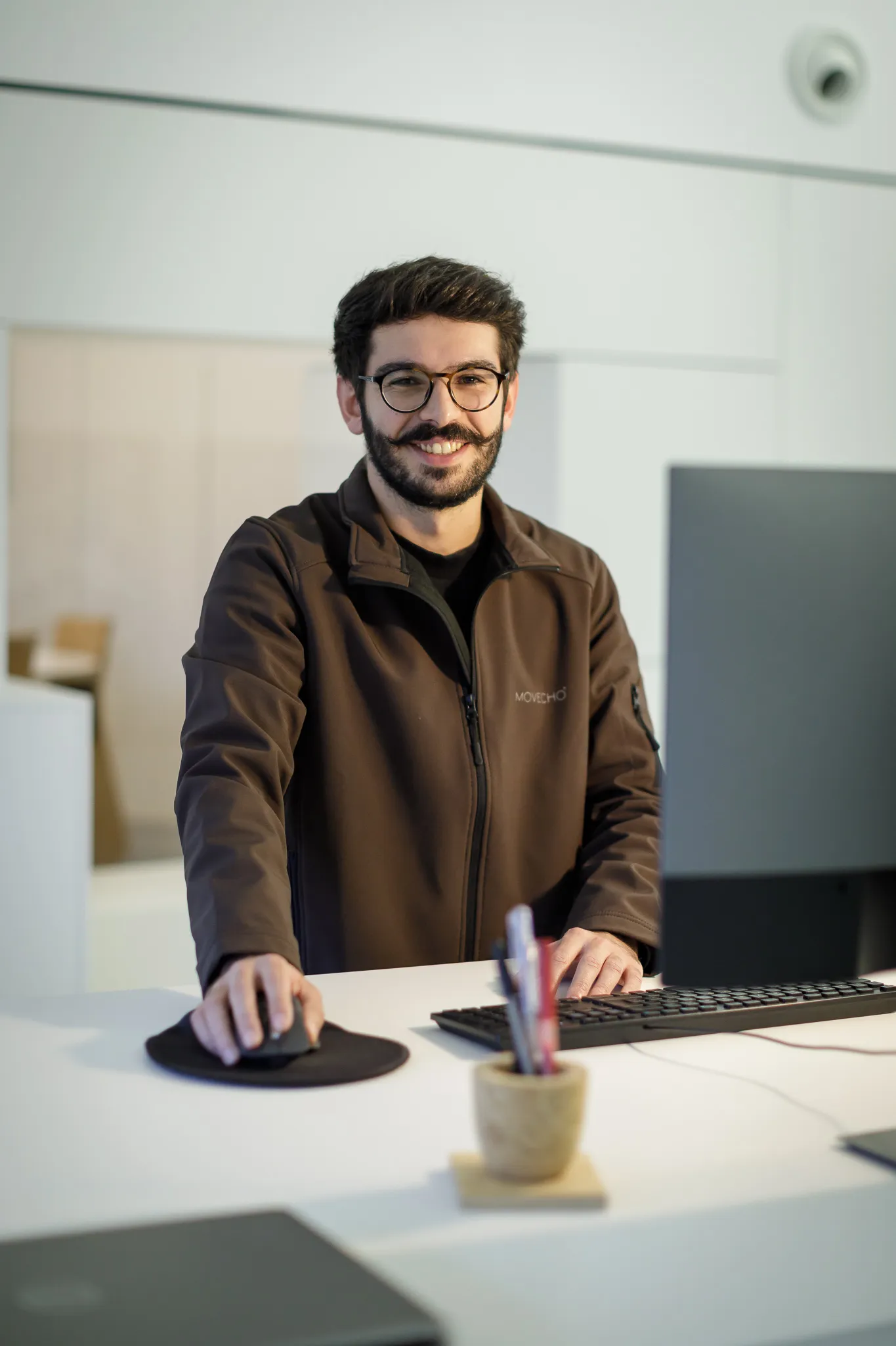 Homem sorridente com óculos usando mouse e teclado em uma mesa de escritório moderna.