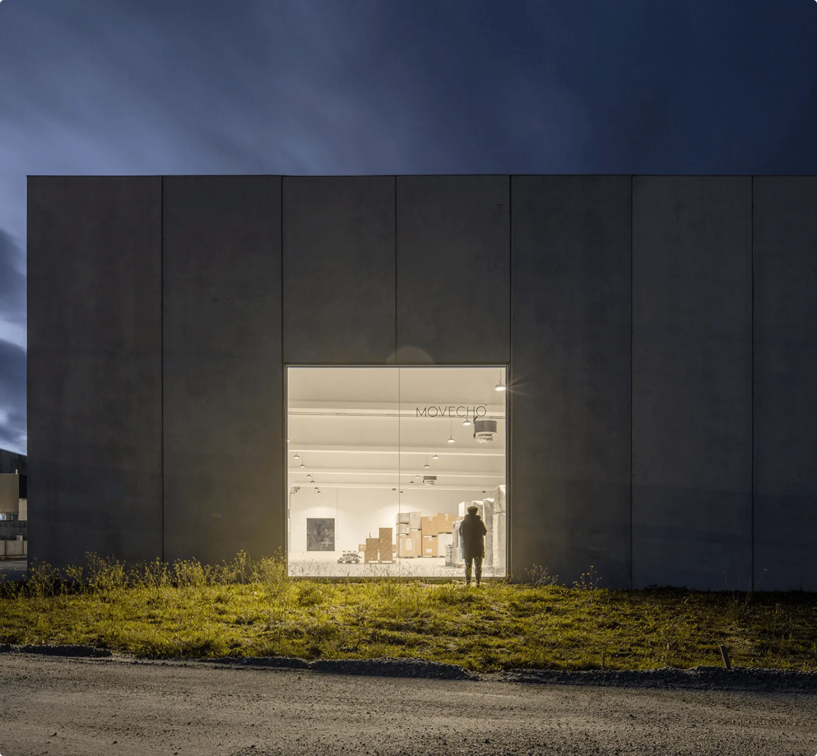 Movecho's factory in Nelas at night: a person outside looks through a large window into the bright interior.