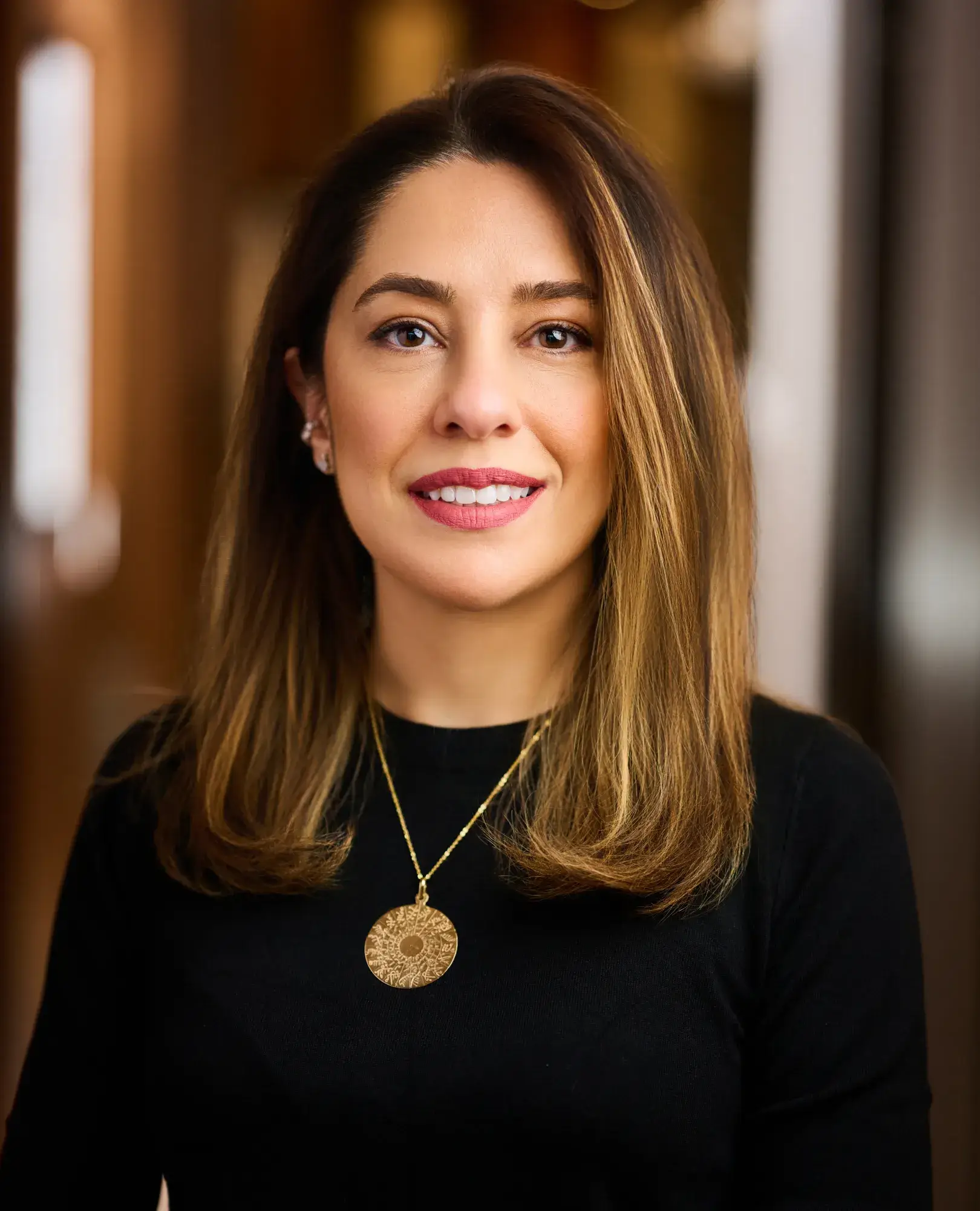 Woman with shoulder-length brown hair wearing a black top and large round gold pendant necklace.