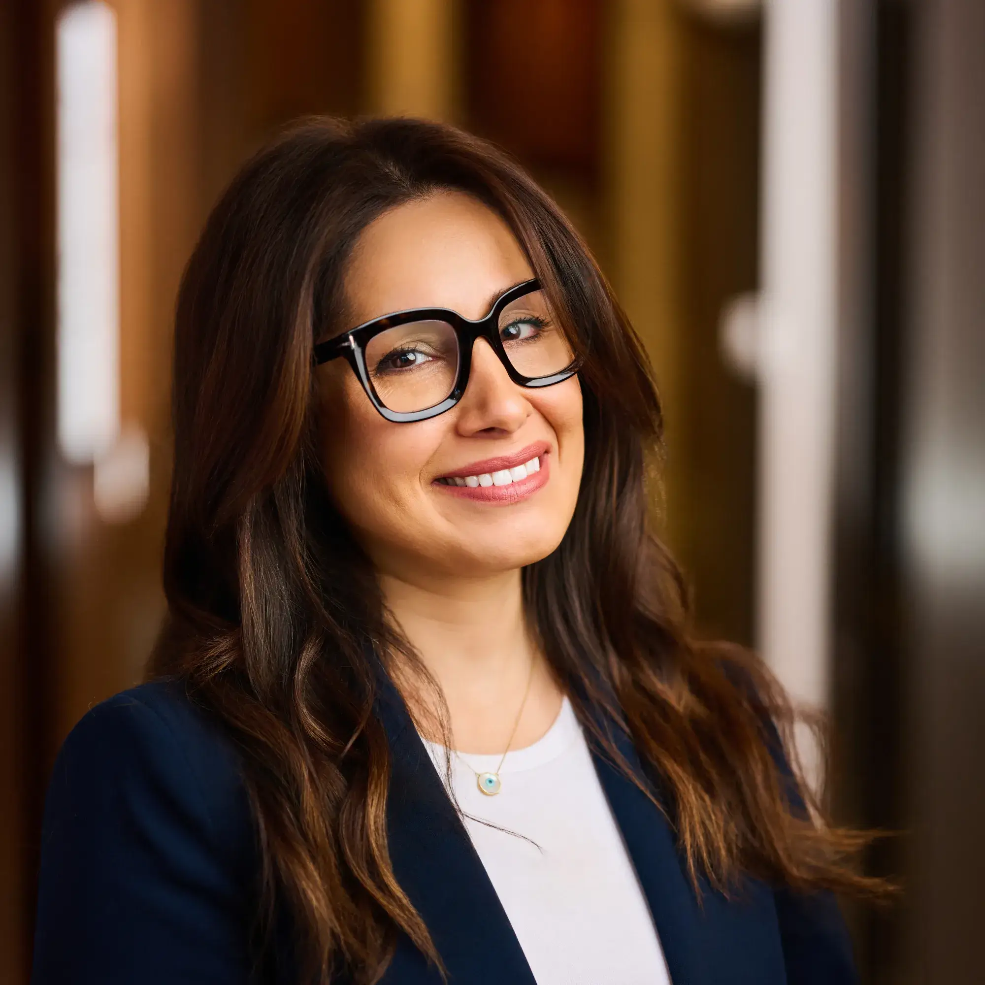 Smiling woman with long brown hair and glasses wearing a navy blazer and white top.