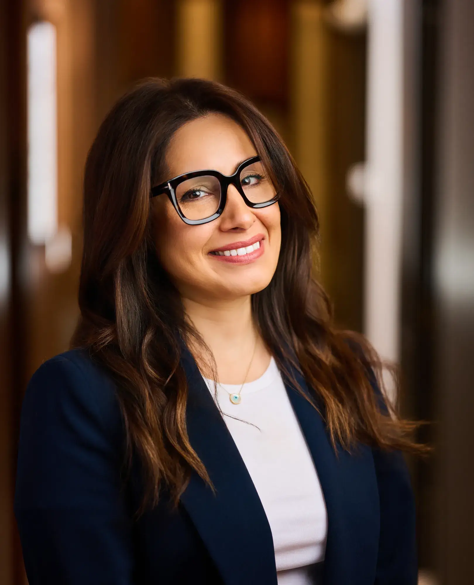 Smiling woman with long brown hair, black glasses, navy blazer, and white top in warm indoor setting.