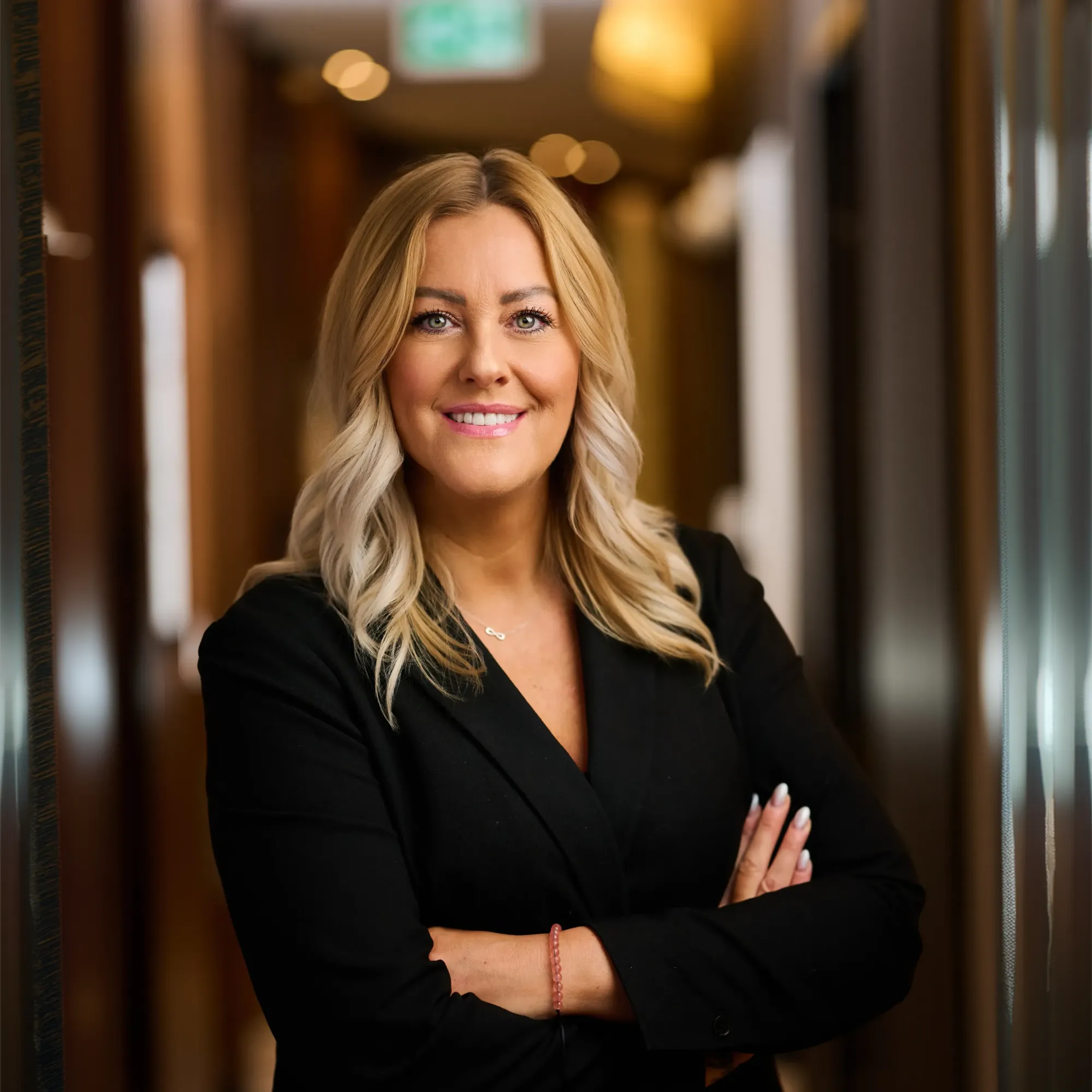 Smiling blonde woman in black jacket with arms crossed in a warmly lit hallway.