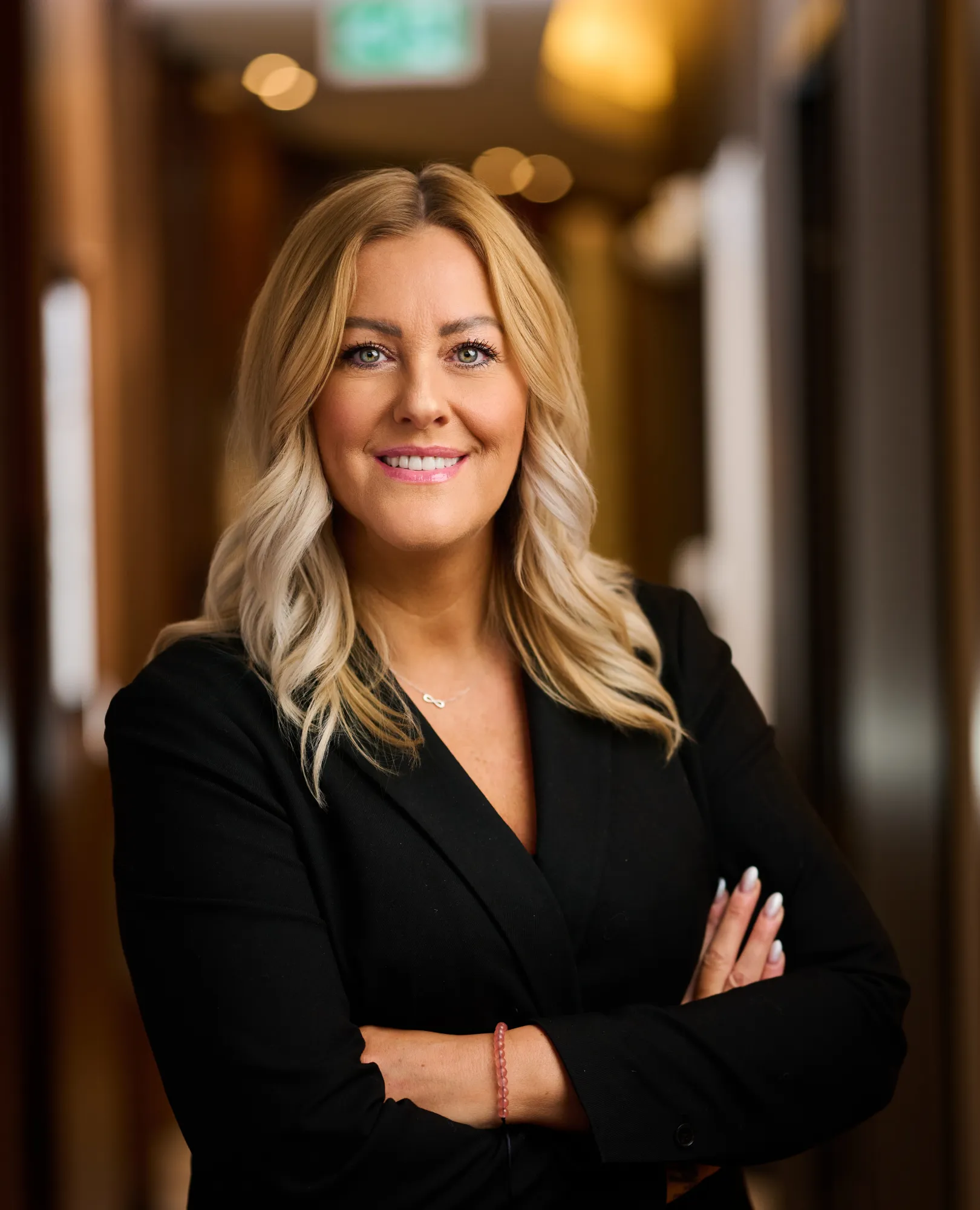 Blonde woman in black blazer with arms crossed smiling in a warmly lit indoor setting.