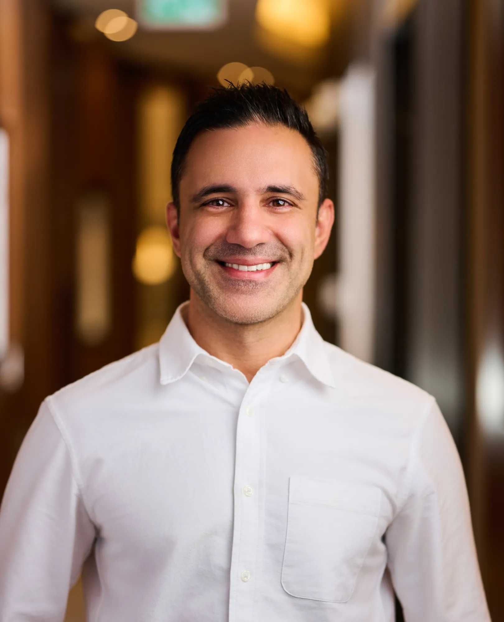 Smiling man with short dark hair wearing a white button-up shirt in a warmly lit hallway.
