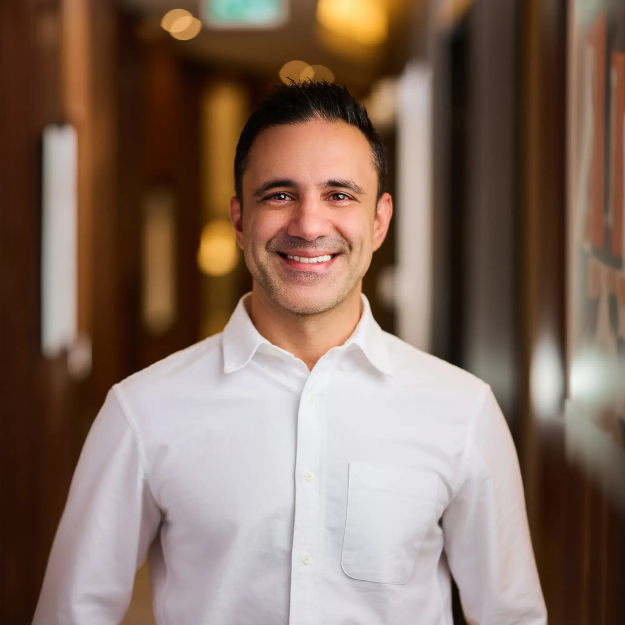 Smiling man with dark hair wearing a white button-up shirt in a warmly lit hallway.