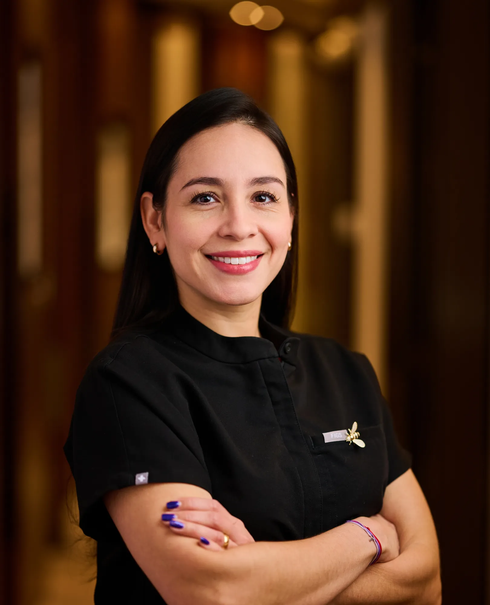 Smiling woman with crossed arms wearing black uniform and blue nail polish in a warmly lit room.
