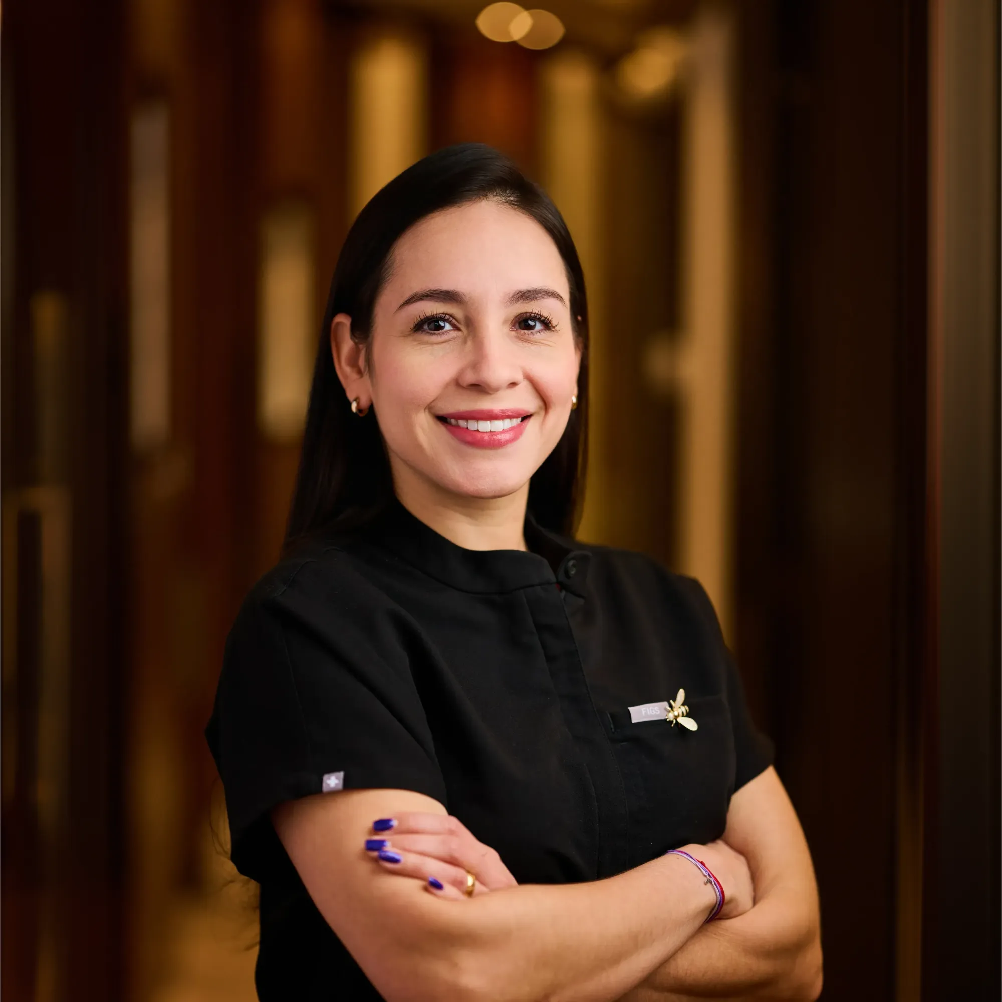 Smiling woman in black medical scrubs with folded arms standing in a hallway.
