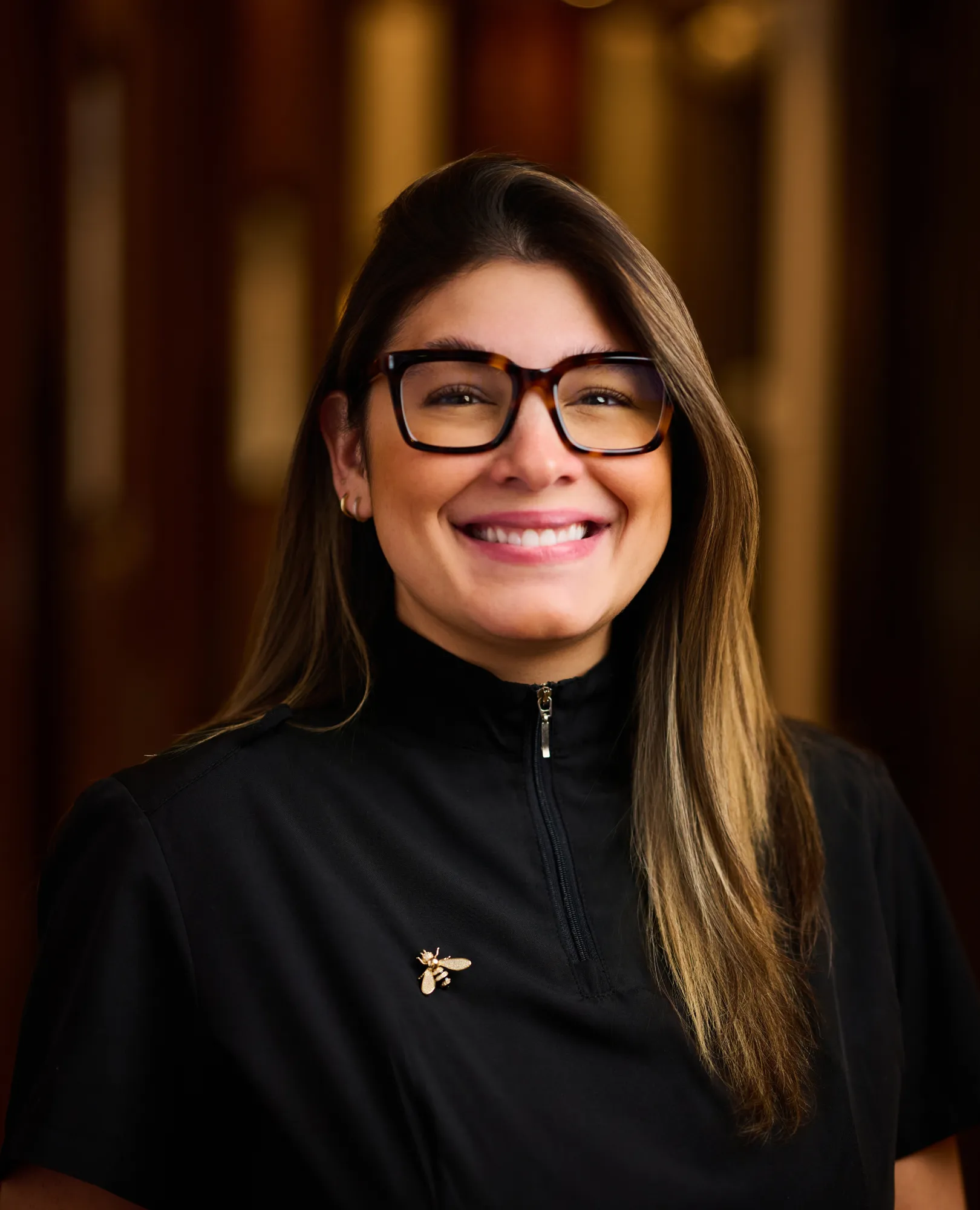 Smiling woman with long hair wearing large glasses and a black zip-up shirt with a bee pin.