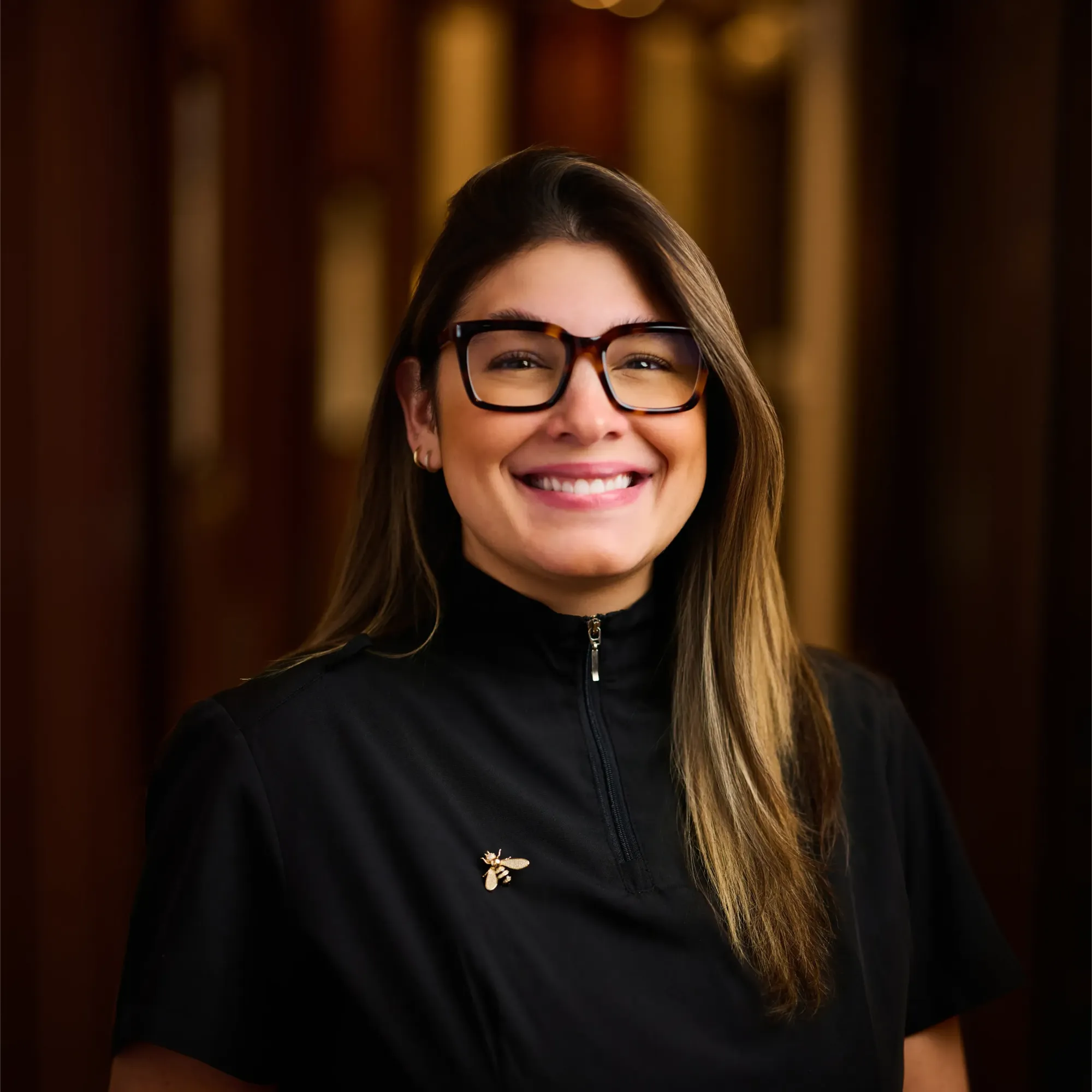 Smiling woman with glasses wearing a black zip-up top with a gold bee pin.
