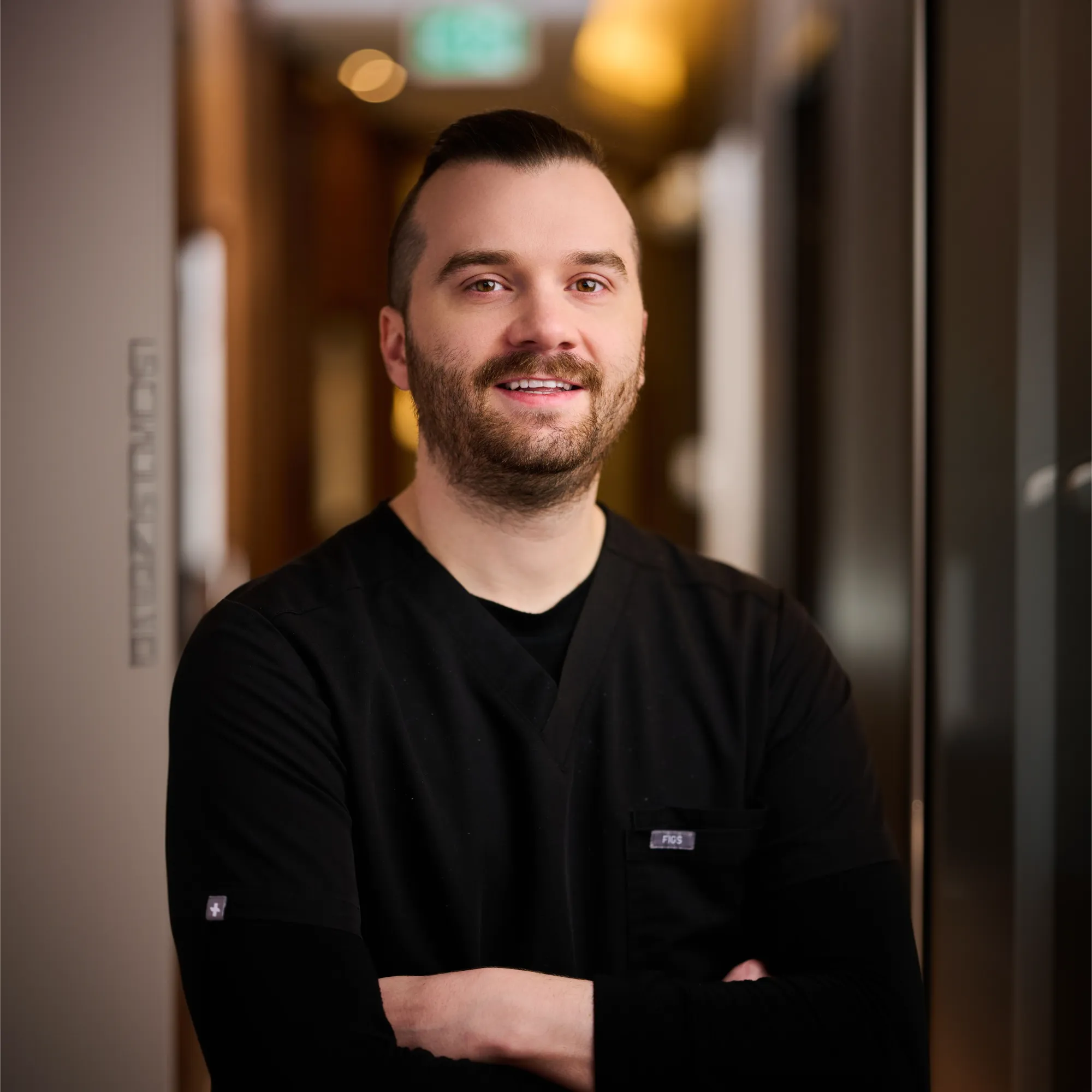 Smiling man with beard wearing black scrubs standing with arms crossed indoors.
