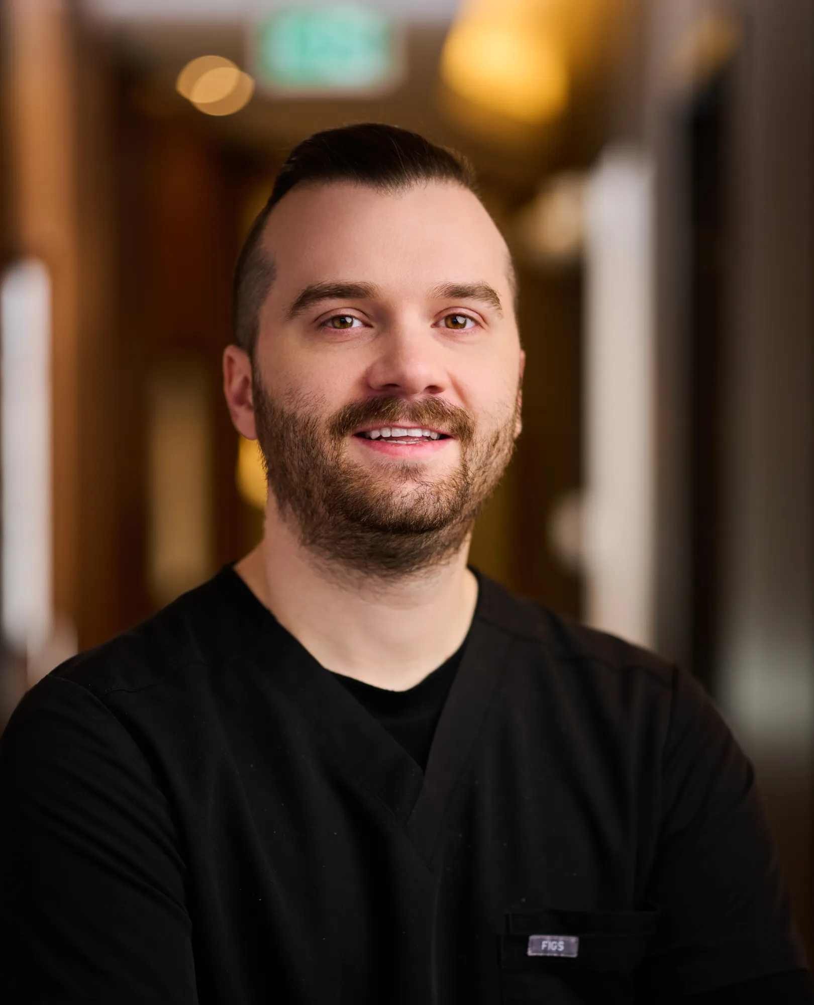 Smiling man with beard wearing black medical scrubs standing indoors in soft light.