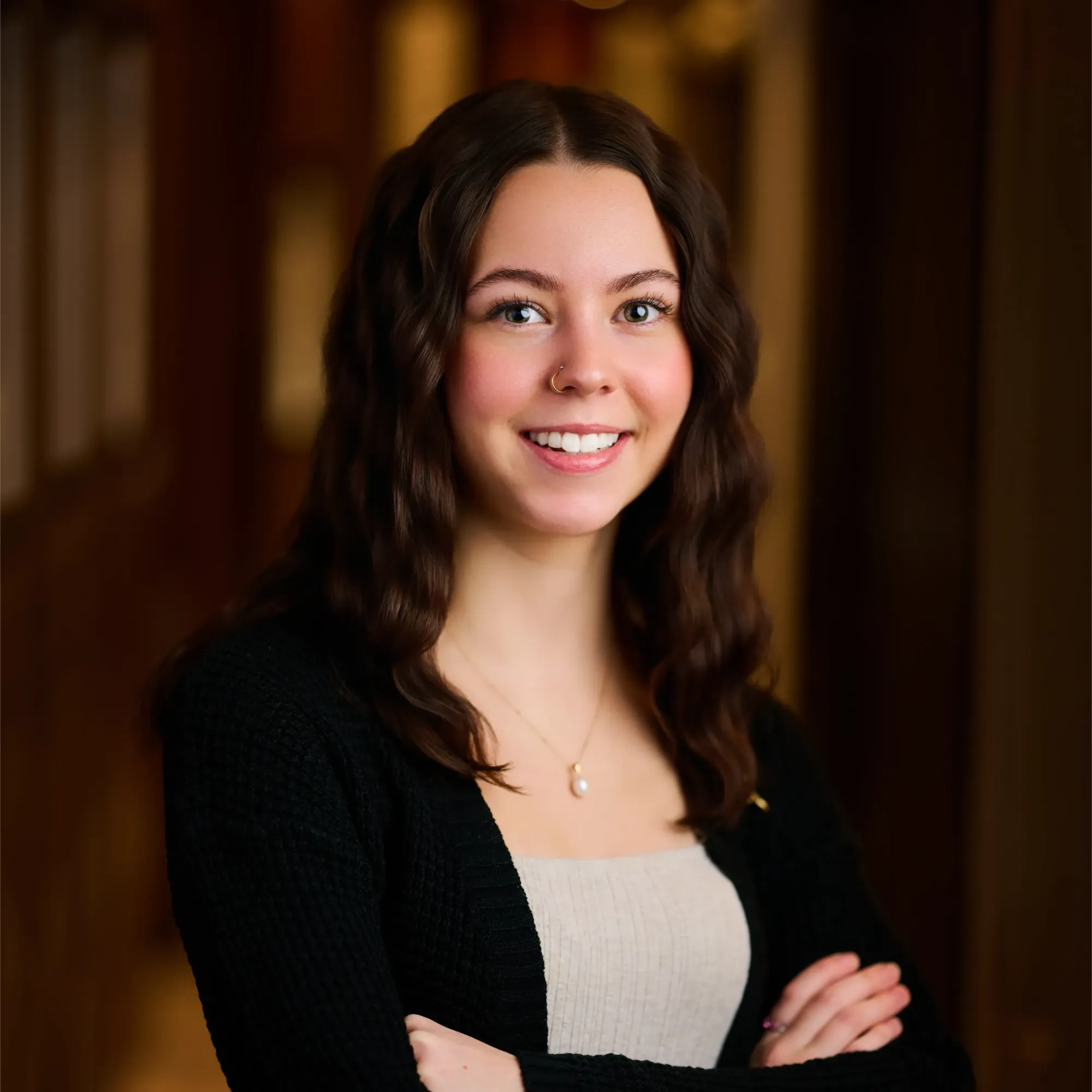 Young woman with wavy brown hair smiling with arms crossed, wearing black cardigan and necklace.