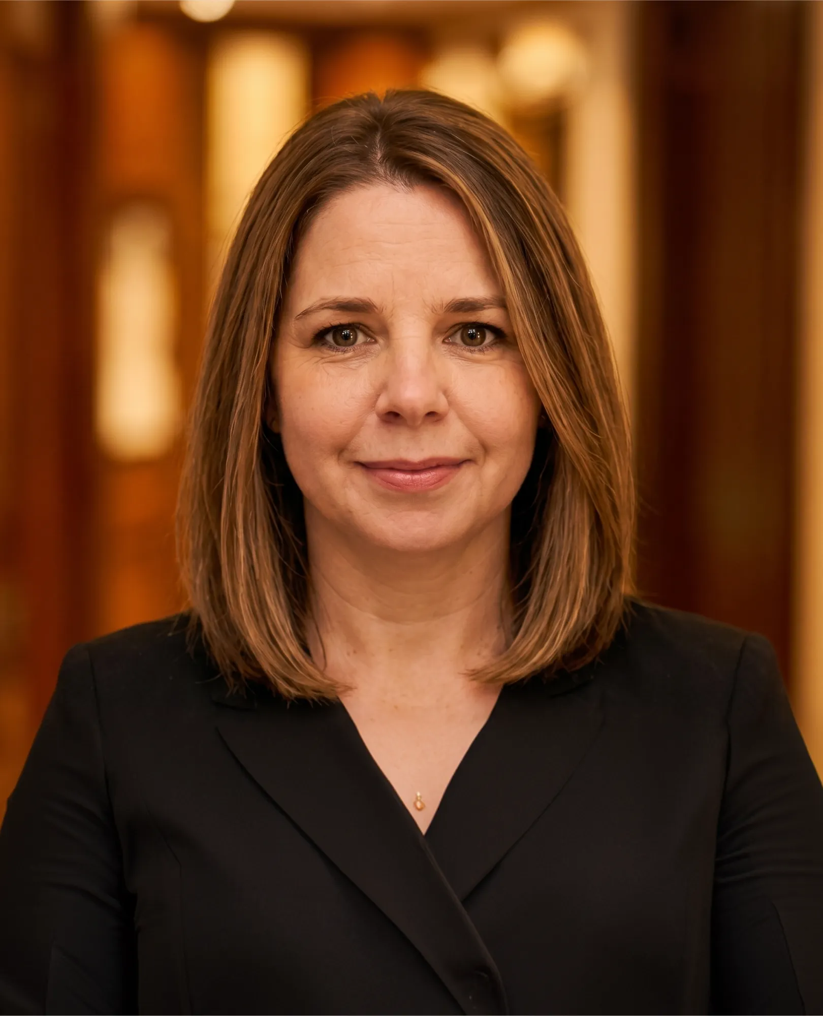 Portrait of a woman with shoulder-length light brown hair wearing a black blazer in a warm interior.