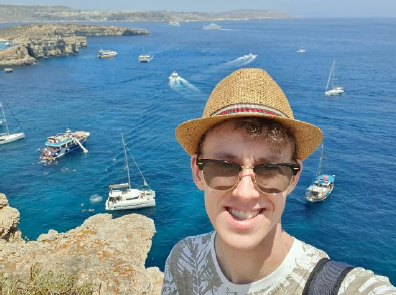 Selfie an der Küste von Malta mit Sonnenhut, im Hintergrund Boote im Meer