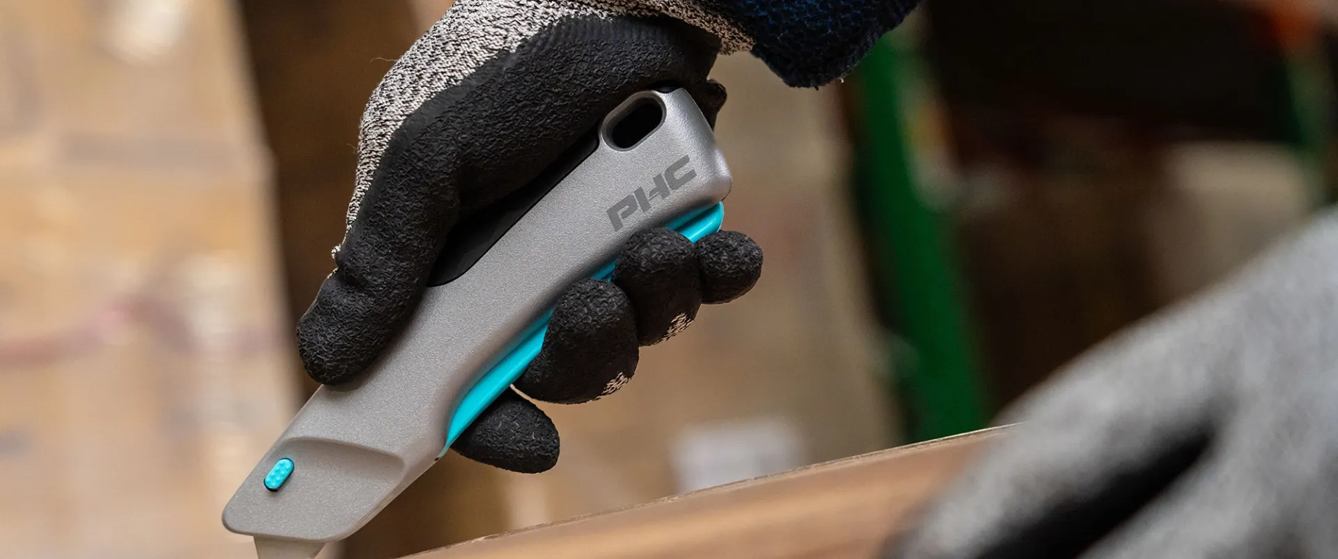 Gloved hand holding a gray utility knife with a blue grip cutting a cardboard box.