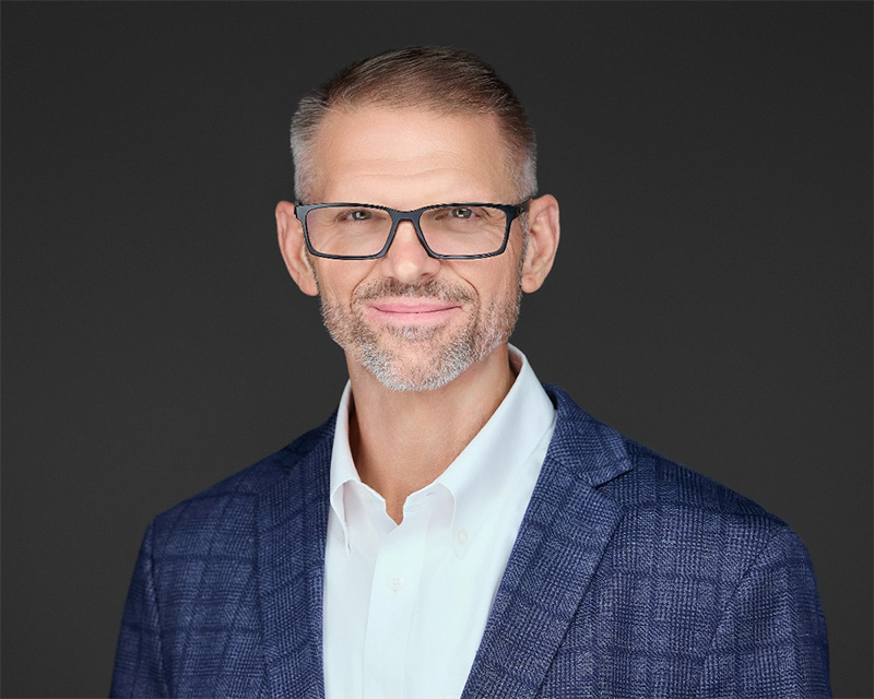 Middle-aged man with short hair, glasses, and a trimmed beard wearing a blue blazer over a white shirt, smiling against a dark background.