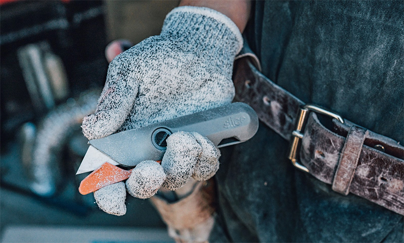 Hand wearing a speckled work glove holding a grey utility knife with an orange safety cover, next to a worn brown leather belt.