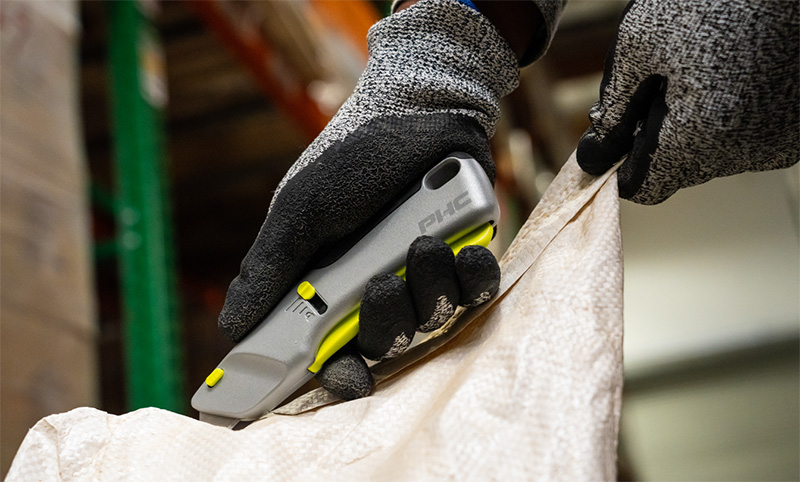 Gloved hands using a gray and yellow utility knife to cut open a white sack in a warehouse.