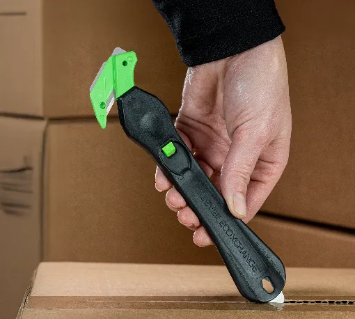 Hand holding a black and green safety box cutter cutting open a cardboard box.
