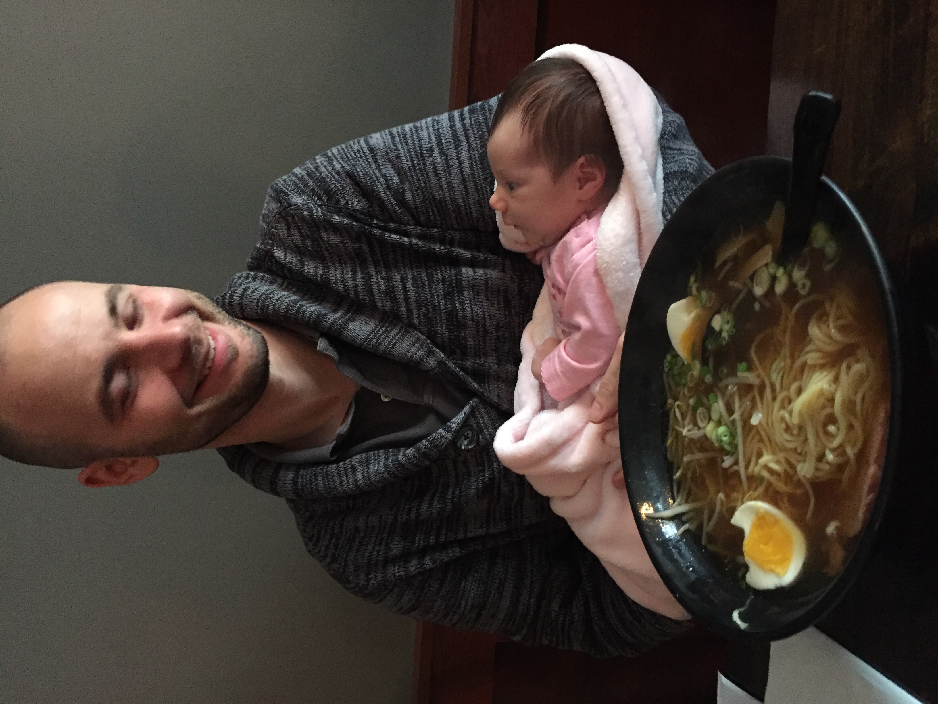 Jesse holding an infant while sitting in front of a bowl of ramen.