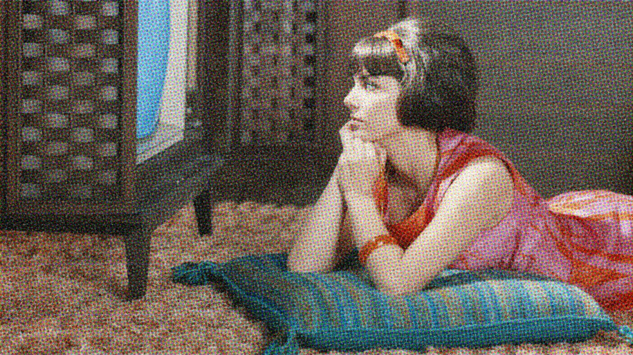 A young woman with a vintage hairstyle lays on a cushion on the floor, gazing at a mid-century TV.