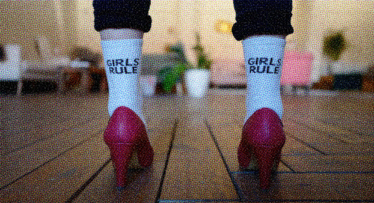 A person wearing dark red high heels and socks stands with their back to us. The shot is close-up on their socks, which ave the words "Girls rule" in black lettering on the back.