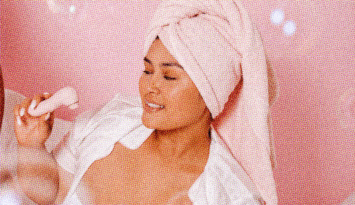 A woman wearing a bath towel on her head holds up a vibrator, smiling.
