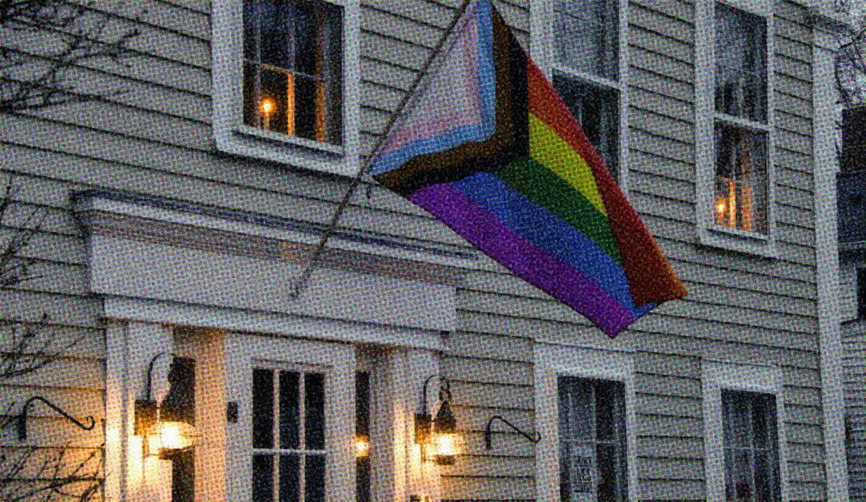 A house flies a Progress Pride Flag on a flag pole mounted over the front door. The family who lives inside welcomes all LGBTQIA+ people and celebrates their identities.