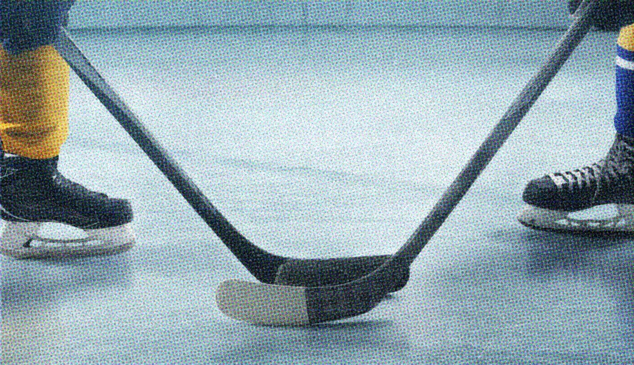 A close-up of two players on an ice hockey rink. We see their skates, sticks, and a puck on the ice between them. They're ready to face off.