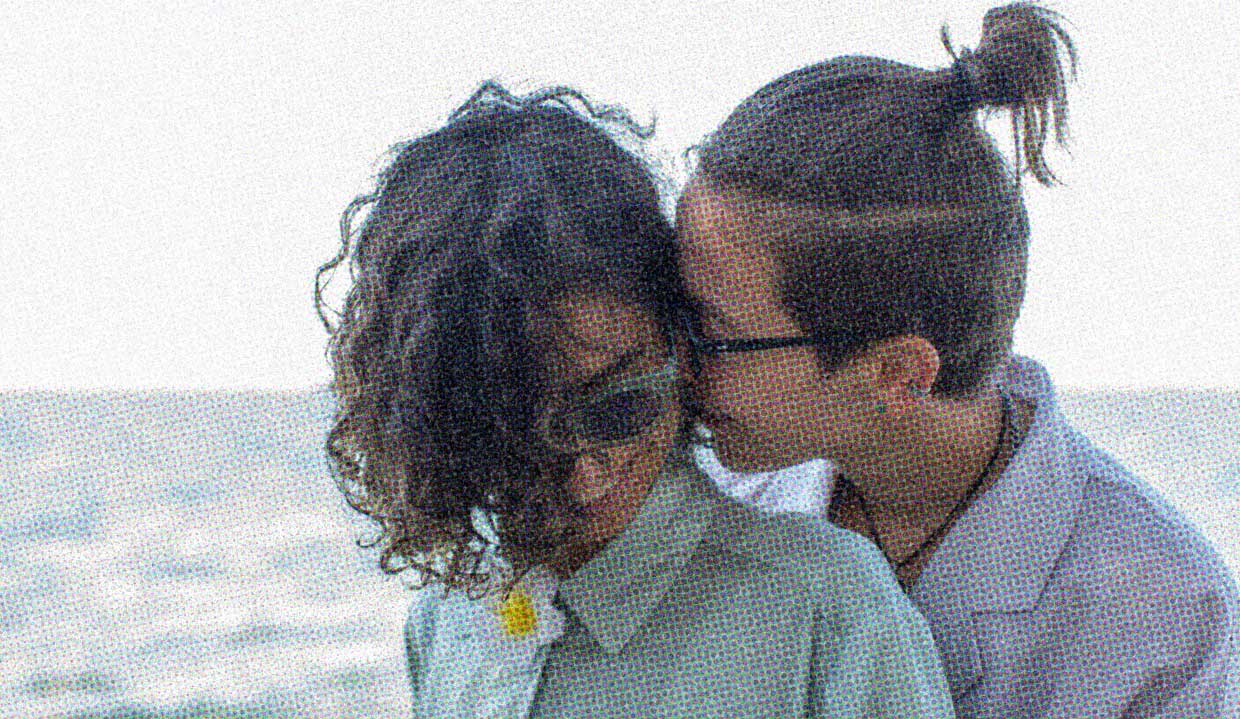 Two people stand close together. One whispers in the ear of the other, who is smiling.