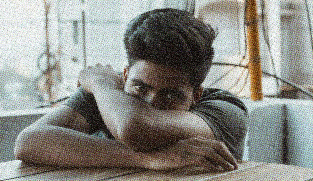 A man sits at a table, his head in his arms, peering over his crossed arms with an expression mixed between hopefulness and despair. He's conflicted about the state of the world, and so are we.