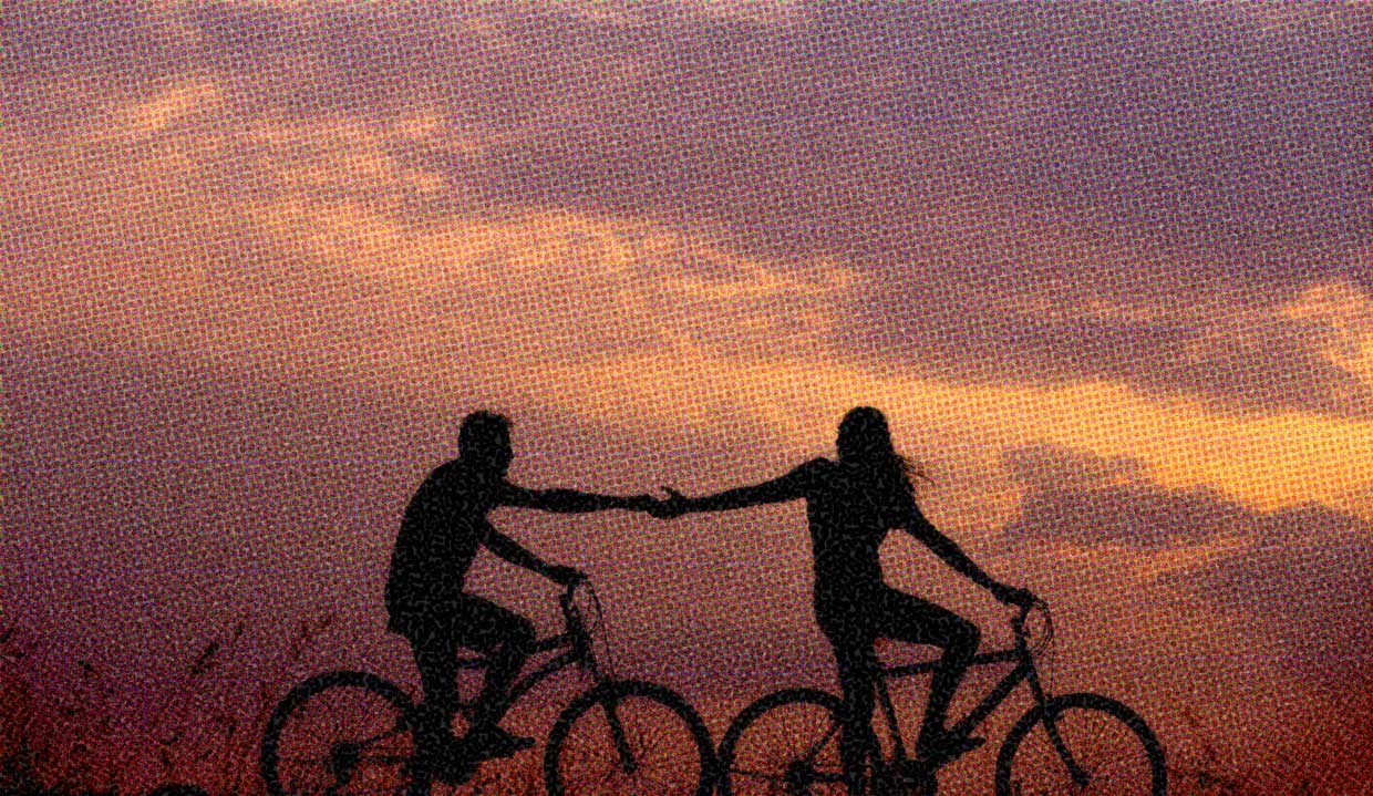 Two people ride bicycles at sunset, silhouetted against the colourful sky. The cyclist in front reaches out a hand for the cyclist behind. Their hands touch.