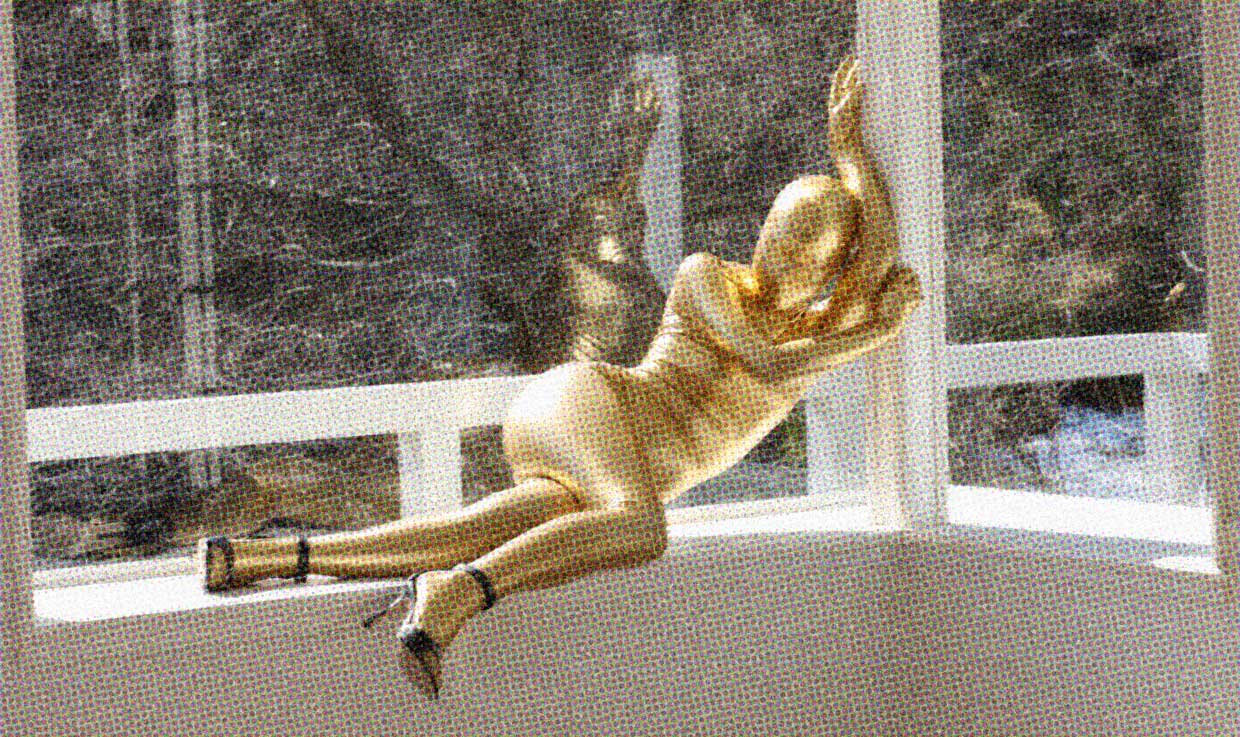 A person wearing a metallic gold zentai suit which covers them from head to toe poses in a window.