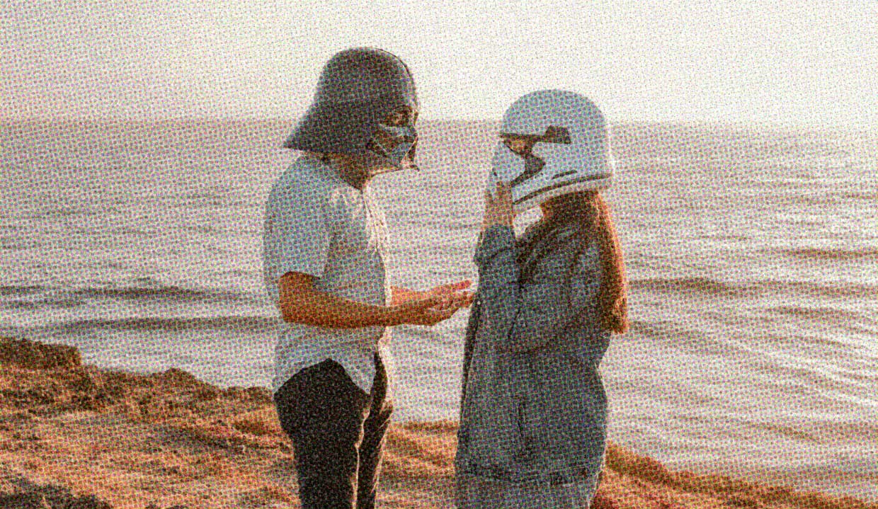 Two people stand on a beach. One is wearing a Darth Vader helmet, while the other is wearing a Stormtrooper helmet. They appear to be in animated conversation.