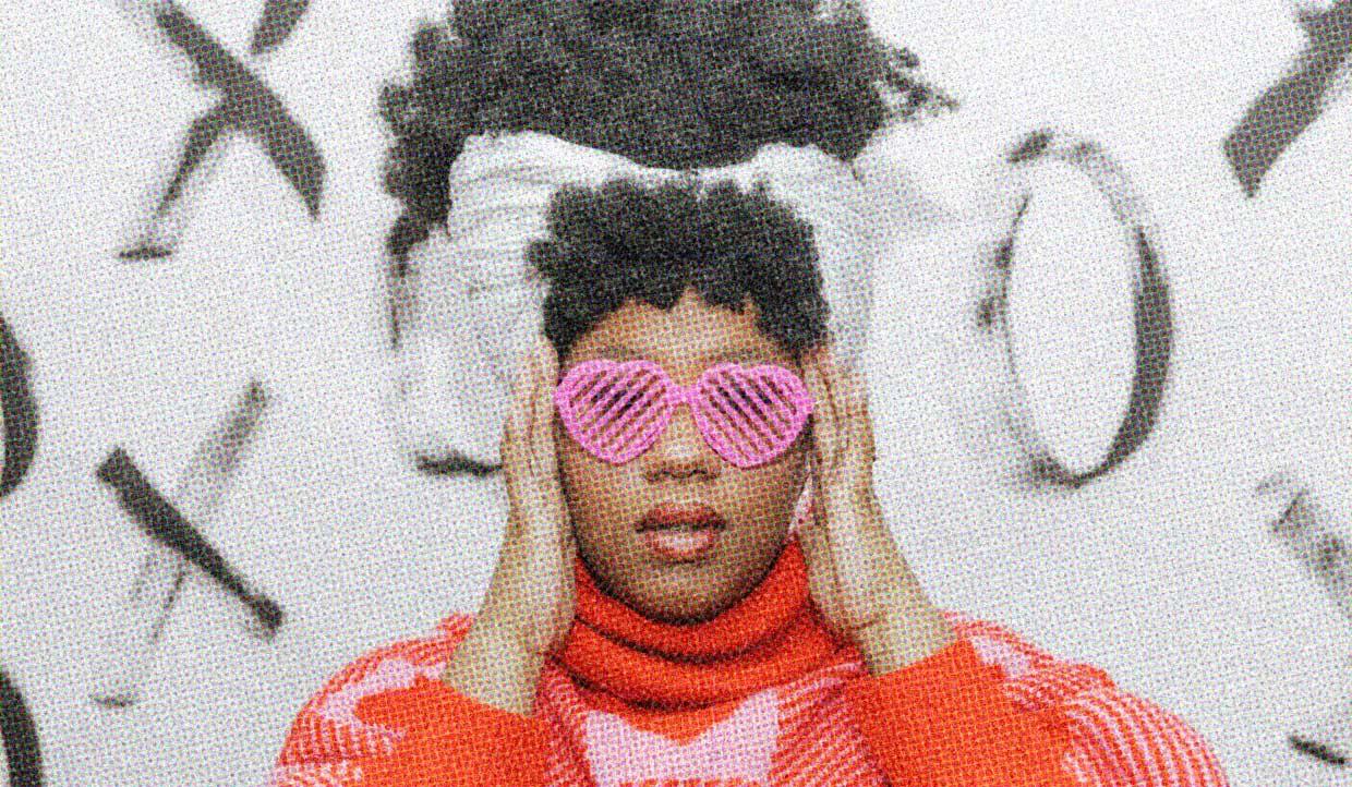 A Black woman wears pink heart-shaped sunglasses and a pink-and-red sweater with hearts printed on it. She stands against a backrdrop with X and O on it.