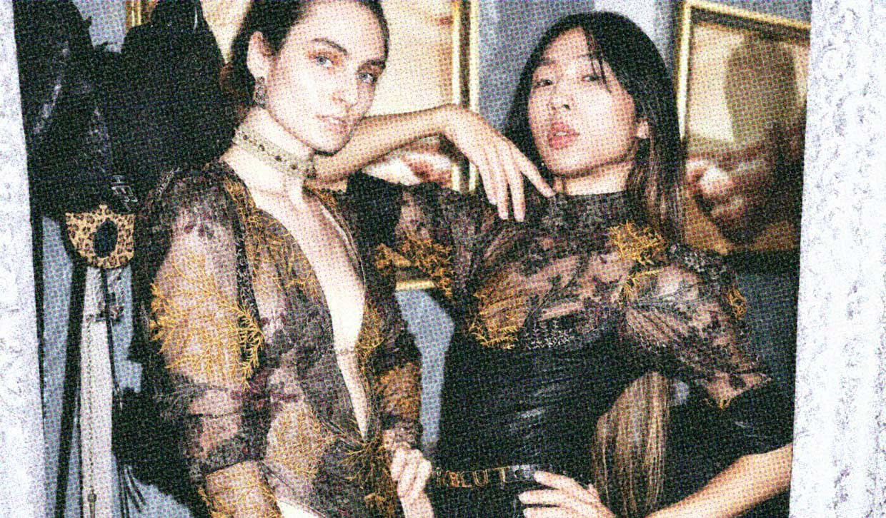Two androgynously features people stand in an elaborately decorated dressing room. They are wearing black haute couture of the sexy variety, with their hair, nails, and makeup done. They look fabulously elegant and sophisticated.