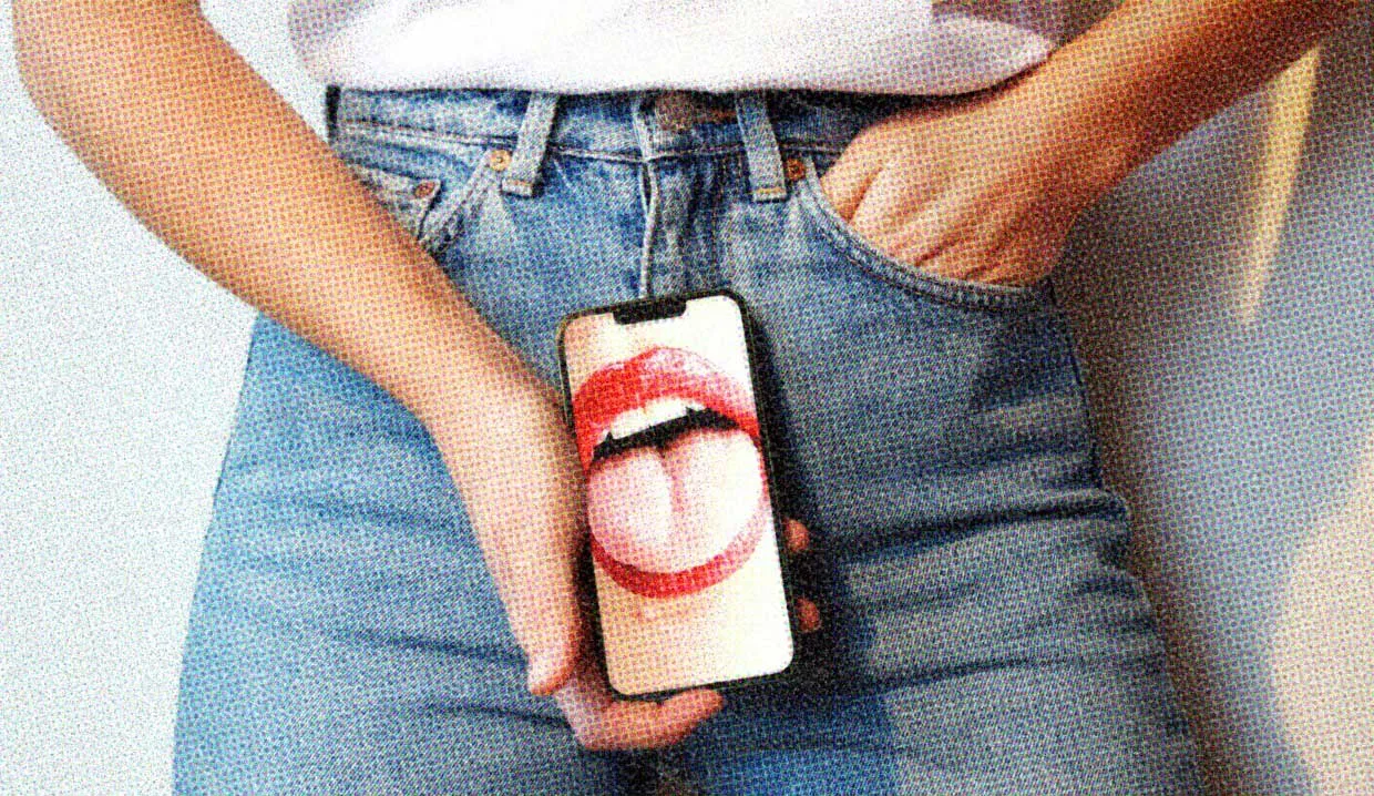 A close-up of a person wearing jeans and holding a mobile phone so the screen is facing the viewer. The phone screen shows a mouth with red lipstick.