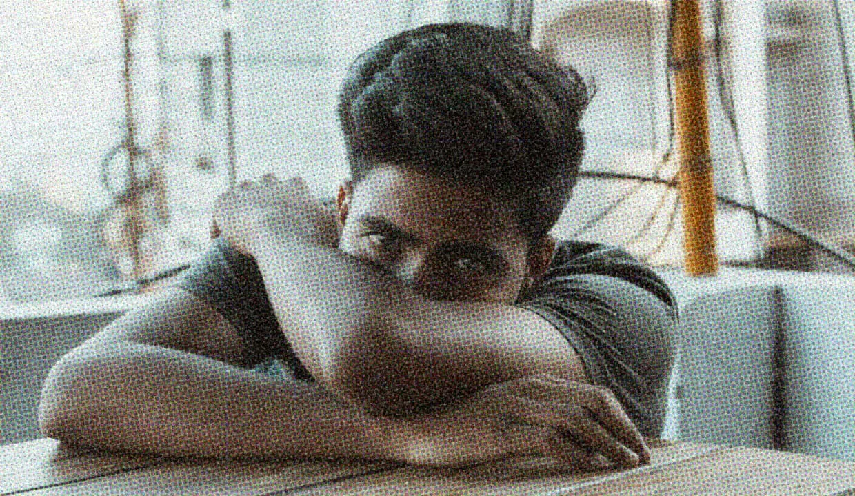 A man sits at a table, his head in his arms, peering over his crossed arms with an expression mixed between hopefulness and despair. He's conflicted about the state of the world, and so are we.