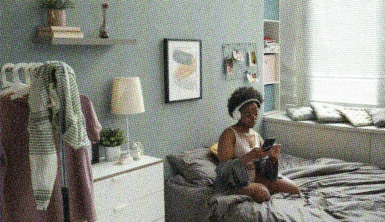 A woman sits on her bed in her bedroom, wearing headphones and vibing. She's not lonely, she's enjoying her alone time in her own space.