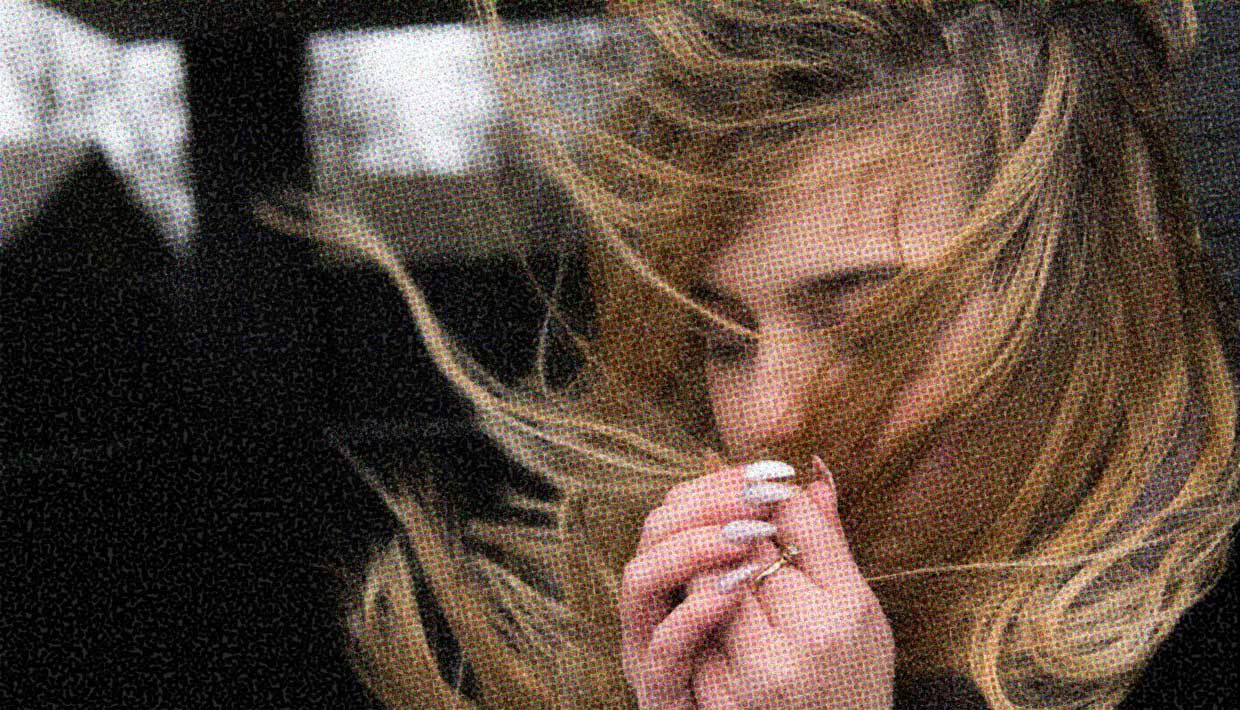 A woman with medium hair stands in a windy spot. Her hair blows around her face. Her hands are raised as though to either protect her hair, or give in to the wind.