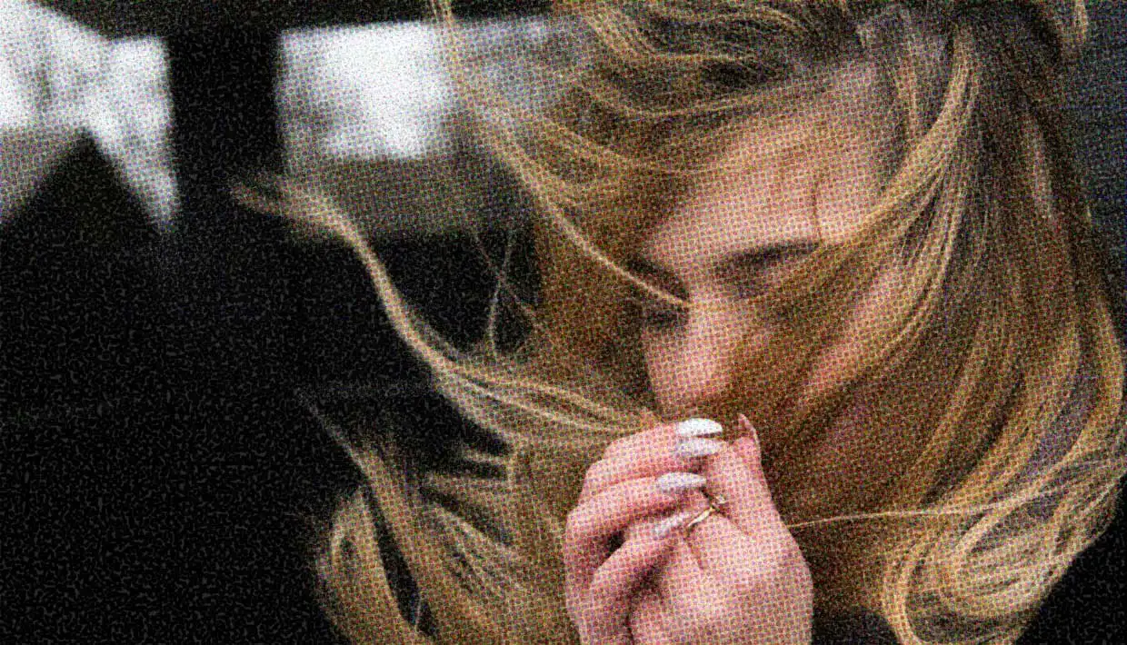 A woman with medium hair stands in a windy spot. Her hair blows around her face. Her hands are raised as though to either protect her hair, or give in to the wind.