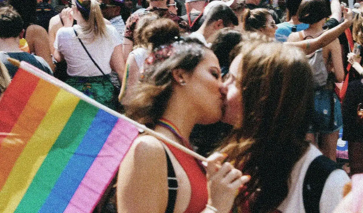 A queer couple kissing at a pride parade, proudly waving a rainbow pride flag.