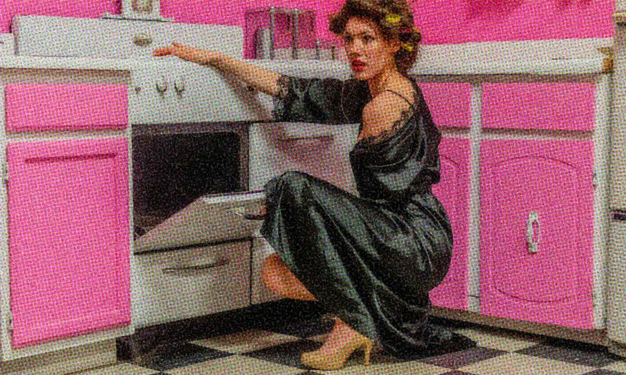 A woman wearing a black satin negligee, with her hair in rollers and with high heels on her feet, bends to look in a cupboard in a pink 1950s-style kitchen.