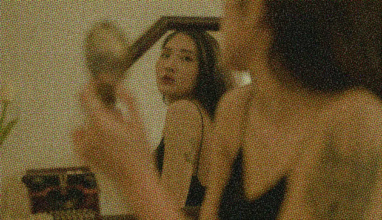 A woman with straight black hair is wearing lingerie and sitting in front of a mirror. She is turned side-on, and is looking at her face in the mirror over her shoulder.