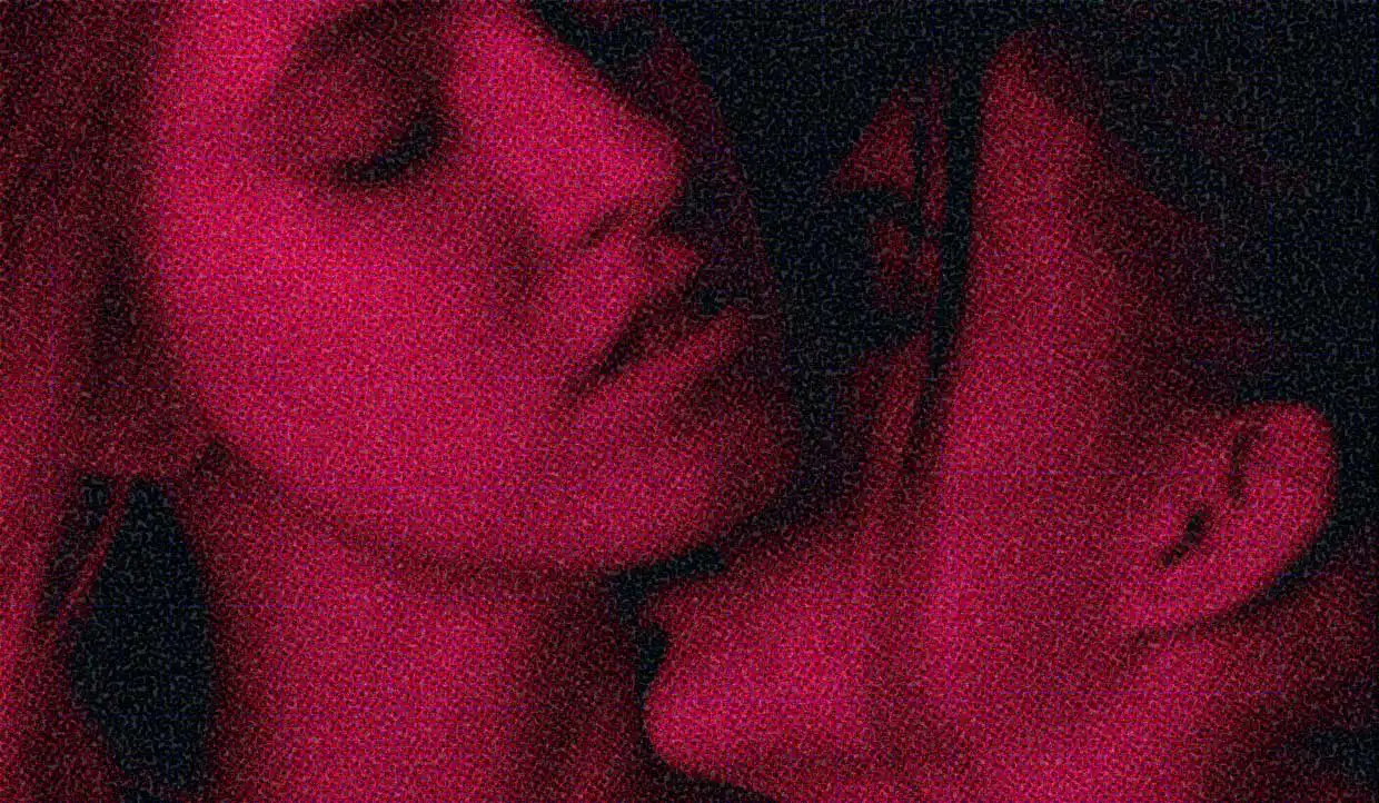 A close-up of two women. One is kissing the other's neck. The lighting is pinky-red.