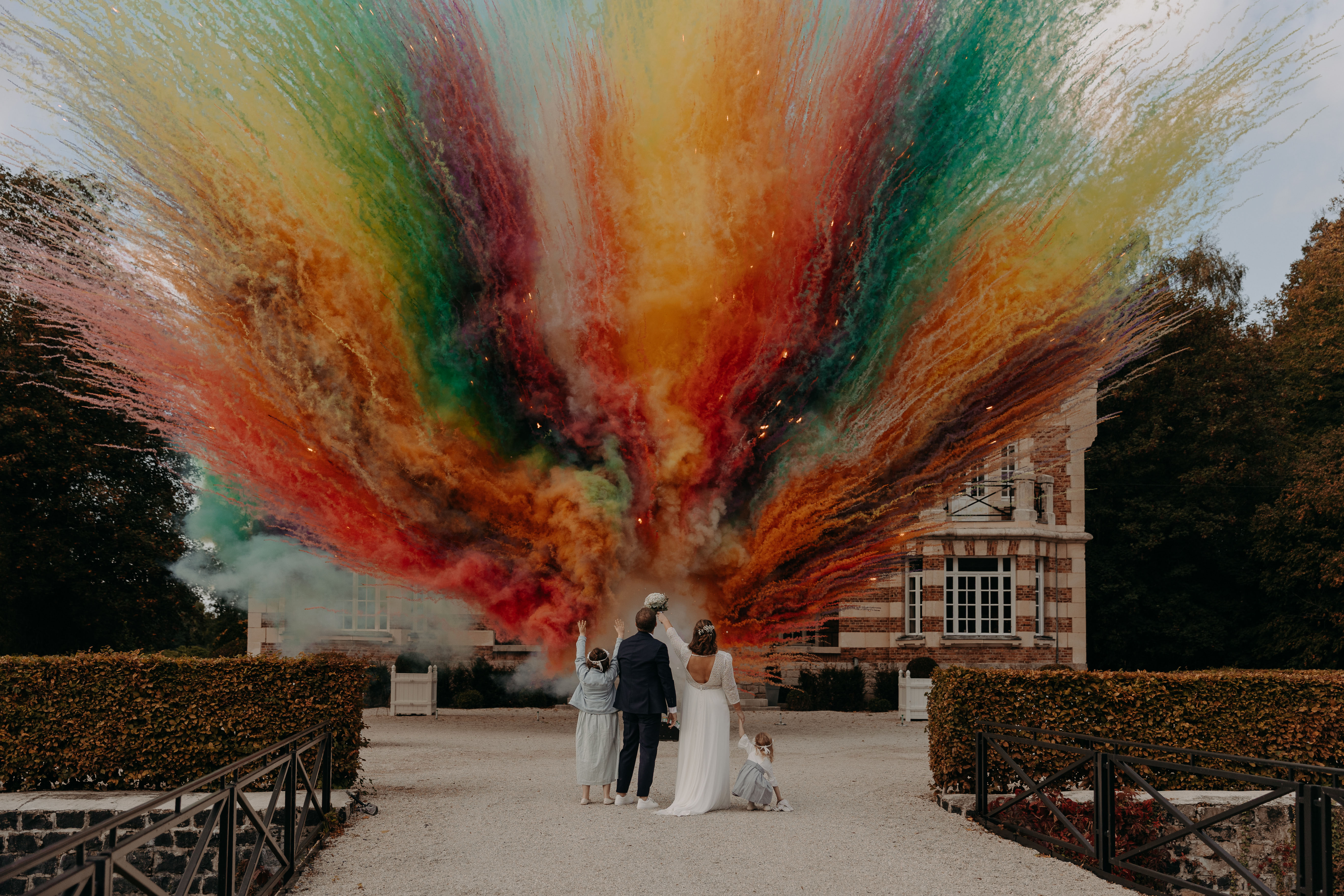 Mariés et leurs enfants admirant une explosion de fumées colorées rouge orange vert et jaune devant un château, feu d'artifice de jour mariage Wagnon Group