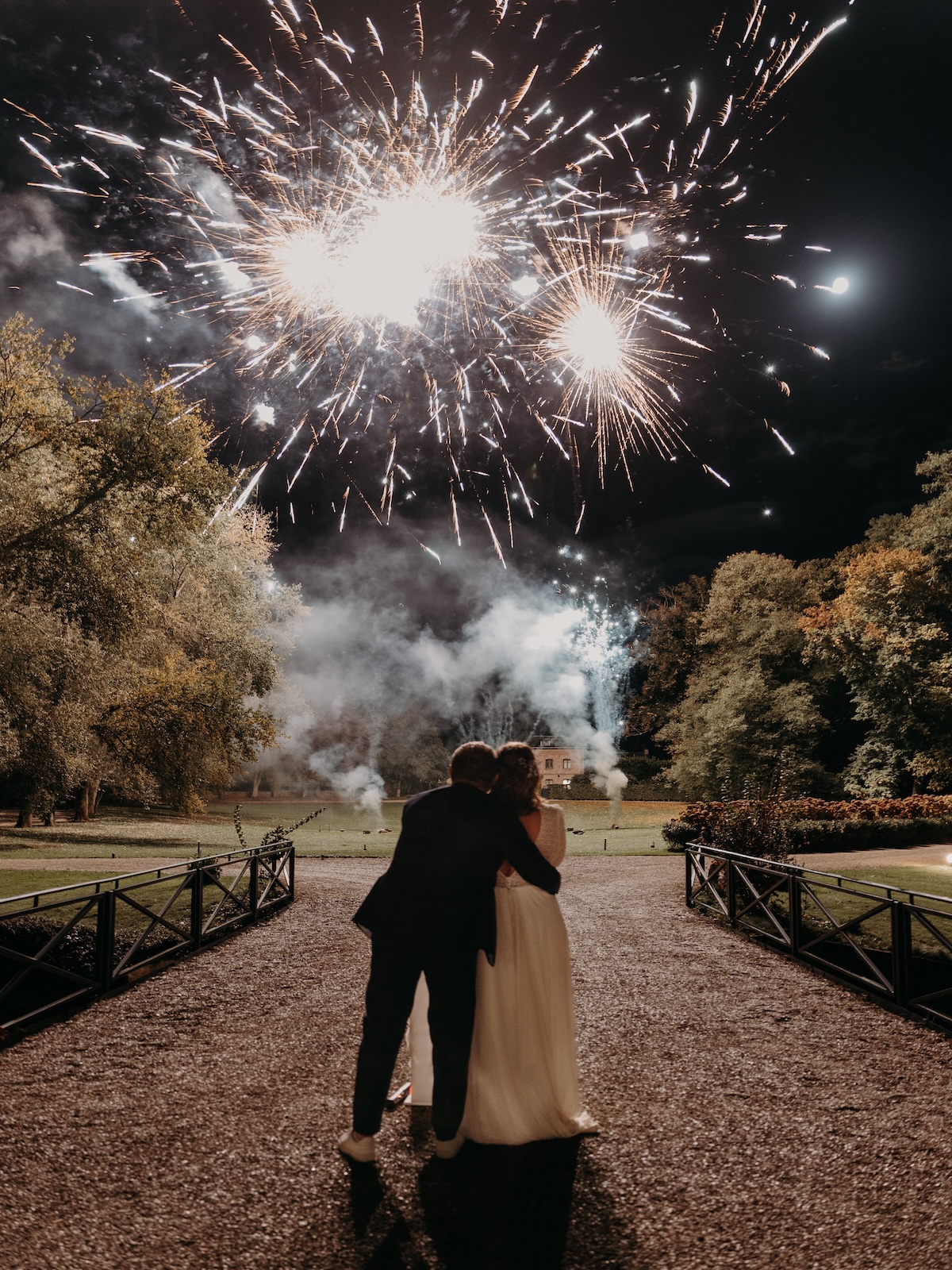 Couple de mariés enlacés admirant un feu d'artifice blanc explosant dans le ciel au-dessus d'un parc, spectacle pyrotechnique Wagnon Group